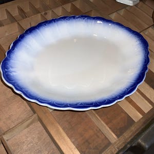 May include: Oval serving platter with a white center and a scalloped edge. The rim features a blue and white gradient design. The platter is made of ceramic and is placed on a wooden surface.