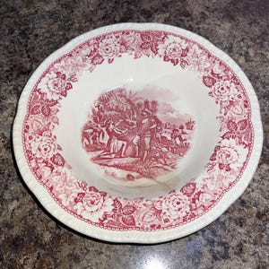 May include: A round ceramic bowl featuring a cream-colored base with a deep red transferware pattern. The rim is decorated with a dense floral motif, while the center depicts a detailed historical scene of figures in a landscape setting.