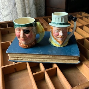 May include: Two ceramic character mugs. One mug is a pirate with an eye patch and a parrot handle. The other mug is a man with a top hat and a handle shaped like a cane. Both mugs are on a blue book.