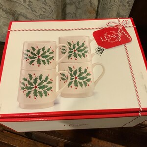 May include: A set of four stacked Lenox mugs in a gift box. The mugs are light pink with a green and red holly design. The box is white with a red border and a red tag that says "Lenox".