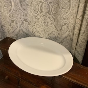 May include: A large, oval, white serving platter. The platter is made of ceramic and has a smooth, glossy finish. The background features a patterned curtain and a dark wooden surface.