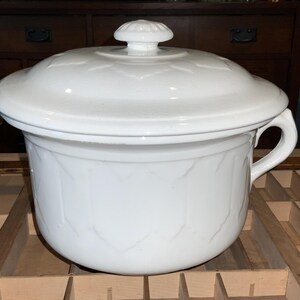 May include: A white ceramic casserole dish with a lid. The dish has a handle and a floral pattern embossed on the sides and lid. The lid has a knob on top. The casserole dish is sitting on a wooden surface.