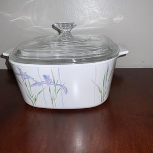 May include: White casserole dish with a clear glass lid and a clear glass handle. The dish is decorated with a floral iris design in shades of purple, blue, and green. The dish is sitting on a dark brown wooden surface.