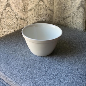 May include: A white ceramic mixing bowl with a smooth, rounded interior and a slightly flared rim. The bowl is sitting on a gray fabric surface.