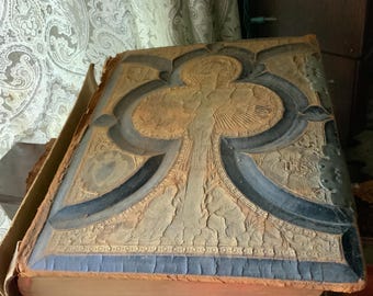 May include: An antique book with a distressed, textured cover. The cover features a detailed embossed design in shades of tan, black, and blue. The book's edges show signs of age and wear, suggesting it is a vintage item.