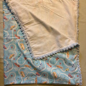 May include: A baby blanket with a light blue car and road pattern. The blanket has a cream-colored interior and a light blue crocheted border. The blanket is approximately 36 inches by 36 inches.