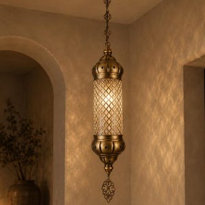 May include: A cylindrical pendant light fixture with an antique brass finish, featuring a central glass section with a diamond-patterned lattice overlay. The lamp hangs from a matching brass chain and includes a decorative metal finial.