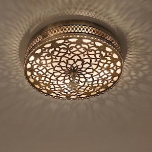 Puede incluir: Lámpara de techo redonda y ornamentada con un diseño calado detallado. La lámpara de metal tiene un acabado dorado antiguo y cálido. La luz proyecta intrincados patrones de sombra en el techo, creando un efecto decorativo.