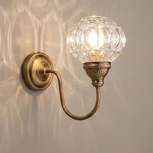 May include: A wall-mounted light fixture with a clear, patterned glass globe and a brass-colored metal base and arm. The light casts a decorative pattern on the wall. The fixture has a vintage or antique aesthetic.