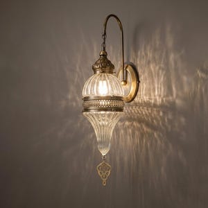May include: A wall-mounted lamp featuring a ribbed, clear glass shade with a teardrop shape. The fixture has an antique brass-toned metal arm and a decorative metal band around the center. A small, ornate metal pendant hangs from the bottom tip.