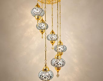 May include: A multi-tiered chandelier featuring six hanging globe-shaped lamps with intricate white and silver mosaic glass patterns. The lamps are suspended from a circular gold-toned metal frame by thin chains, creating a cascading arrangement.
