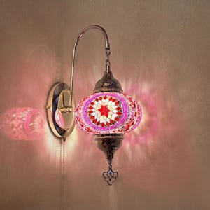 Op de afbeelding: Een decoratieve wandlamp met een mozaïek glazen kap in roze, rood en wit. De lamp heeft een zilverkleurig metalen frame en een trekketting. Het licht werpt een roze gloed op de muur.