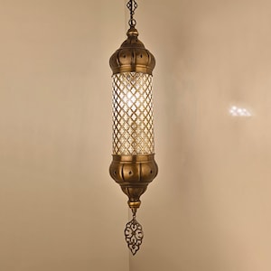 May include: A cylindrical pendant light fixture with an antique brass finish. The central section features a clear glass cylinder encased in a decorative brass lattice grid. It hangs from a matching brass ceiling canopy by a chain.