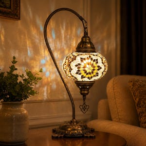 May include: A table lamp with a curved bronze-toned metal stand and a mosaic glass shade featuring a star-shaped pattern in shades of yellow, brown, and white. The lamp emits a warm glow, casting intricate light patterns on the wall behind it.