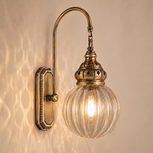 May include: A wall-mounted light fixture featuring an antique brass finish, a rectangular backplate, a curved arm, and a decorative chain link. The light includes a clear, ribbed glass globe shade with a metal cap containing small diamond cutouts.