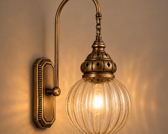 May include: A wall-mounted light fixture featuring an antique brass finish, a rectangular backplate, and a curved arm. A spherical, ribbed clear glass shade hangs from a chain, topped with a decorative brass cap with diamond cutouts.