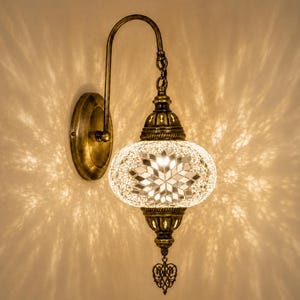May include: A wall-mounted lamp featuring a brass-toned metal fixture and a spherical shade crafted from white and clear glass mosaic tiles. The lamp emits a warm, patterned light, and includes a decorative metal finial hanging from the base.