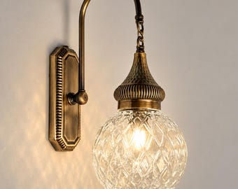 May include: A wall-mounted light fixture featuring an antique brass finish, a curved arm, and a rectangular backplate. The lamp includes a textured, bell-shaped metal cap and a spherical, clear glass shade with a diamond-cut pattern.