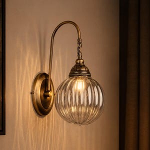 Puede incluir: Una lámpara de pared con un brazo curvo de latón antiguo y una pantalla de globo de cristal transparente con estrías verticales. El accesorio incluye una tapa decorativa de latón en la parte superior del globo, emitiendo una luz cálida.
