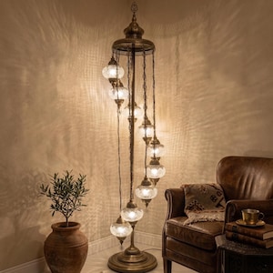 Op de afbeelding: Een sierlijke vloerlamp met een bronzen afwerking. De lamp heeft meerdere glazen bolvormige lampenkappen die in een spiraalvorm zijn gerangschikt. De lamp staat naast een bruine leren fauteuil en een potplant.