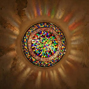 May include: A circular mosaic lamp with a vibrant star pattern. The lamp is composed of numerous coloured glass pieces, including red, green, blue, and yellow. It is set against a textured brown background, with light beams emanating outwards.