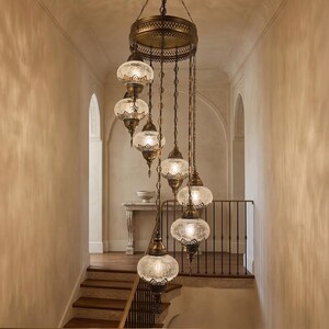 May include: An ornate, multi-tiered chandelier with glass globe shades hangs in a stairwell. The fixture has a bronze finish and intricate detailing. The stairwell features wooden steps, a wrought iron railing, and a neutral color palette.