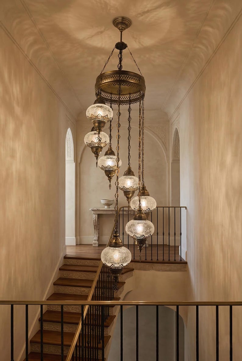May include: An ornate, multi-tiered chandelier with glass globe shades hangs in a stairwell. The fixture has a bronze finish and intricate detailing. The stairwell features wooden steps, a wrought iron railing, and a neutral color palette.