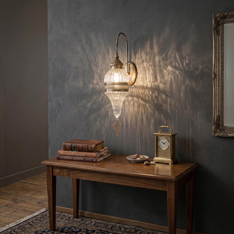 May include: A wall-mounted lamp featuring a teardrop-shaped, ribbed glass shade with antique brass metal accents. The fixture emits a warm light, casting intricate, patterned shadows against the wall. A decorative metal finial hangs from the base.