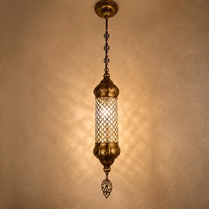 May include: A cylindrical pendant light fixture featuring an antique brass finish, a central glass section with a decorative metal lattice overlay, and a pointed finial at the bottom, suspended from a matching brass ceiling canopy.