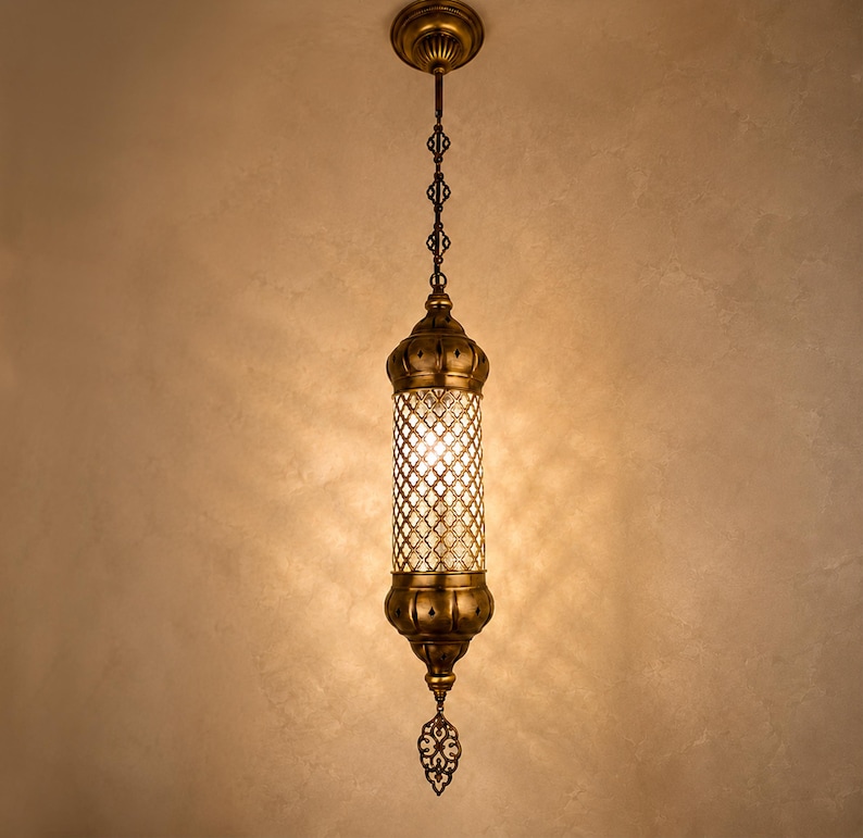 May include: A cylindrical pendant light fixture featuring an antique brass finish, a central glass section with a decorative metal lattice overlay, and a pointed finial at the bottom, suspended from a matching brass ceiling canopy.