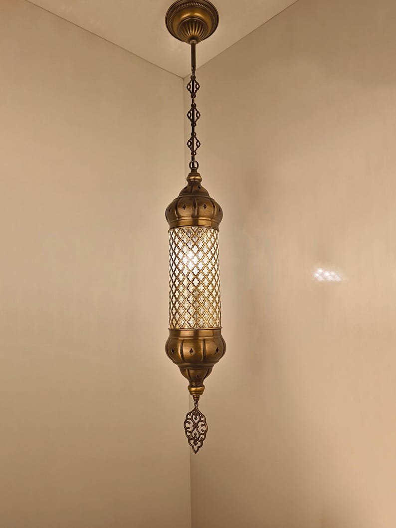 May include: A cylindrical pendant light fixture with an antique brass finish. The central section features a clear glass cylinder encased in a decorative brass lattice grid. It hangs from a matching brass ceiling canopy by a chain.