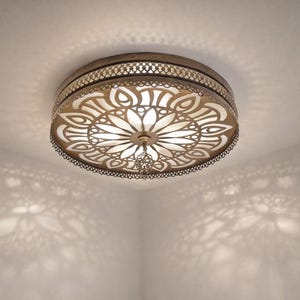 May include: Circular ceiling light with a floral design, made of metal with a bronze finish. The light casts decorative shadows on the white ceiling and walls. The fixture has an intricate, openwork pattern, creating a beautiful effect.
