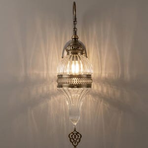 May include: A wall-mounted lamp featuring a clear, vertically ribbed glass shade with a teardrop shape. The fixture includes antique brass-toned metal accents, a central decorative band, and a dangling metal ornament at the bottom.