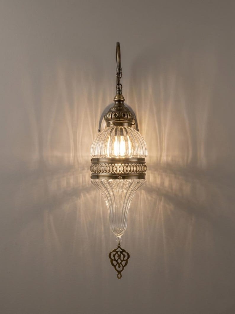 May include: A wall-mounted lamp featuring a clear, vertically ribbed glass shade with a teardrop shape. The fixture includes antique brass-toned metal accents, a central decorative band, and a dangling metal ornament at the bottom.