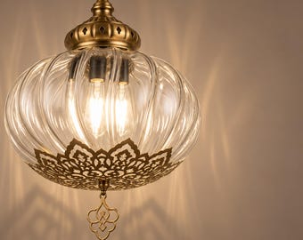 May include: A pendant light fixture featuring a ribbed, clear glass globe shade with an antique brass finish metal cap and base. The base includes an intricate, laser-cut floral metal overlay and a decorative dangling metal finial.