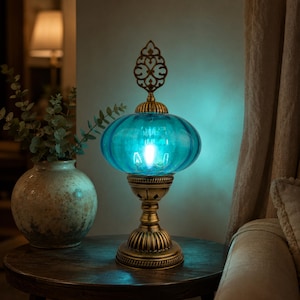 Peut inclure: Une lampe de table dotée d'un abat-jour en verre turquoise translucide arrondi et d'une base en métal couleur laiton antique. La lampe est surmontée d'un fleuron décoratif en métal orné. La lumière émet une lueur douce et froide.