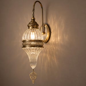 May include: A wall-mounted lamp featuring a brass-toned metal fixture and a clear, ribbed glass shade. The lamp has an ornate, teardrop-shaped design with a decorative metal finial hanging from the bottom and a curved arm mount.