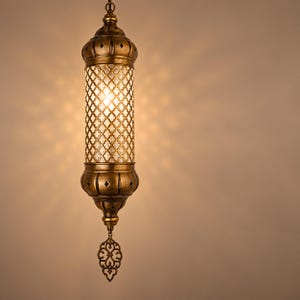 May include: A cylindrical hanging pendant lamp with an antique brass finish. The central section features a clear glass panel with a gold-toned lattice overlay. The top and bottom caps have cut-out diamond shapes, and a decorative metal finial hangs below.