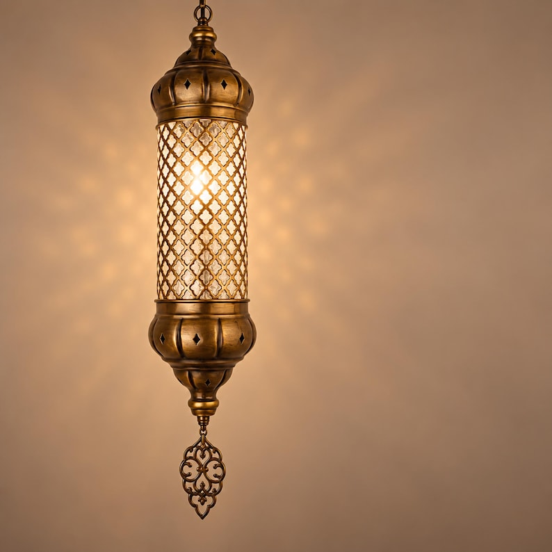 May include: A cylindrical hanging pendant lamp with an antique brass finish. The central section features a clear glass panel with a gold-toned lattice overlay. The top and bottom caps have cut-out diamond shapes, and a decorative metal finial hangs below.