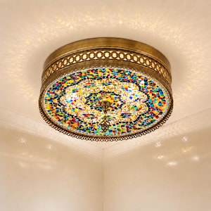 Puede incluir: Una lámpara de techo con una pantalla de mosaico de vidrio multicolor, incluyendo rojo, azul, amarillo y verde. La lámpara tiene un marco de metal color latón con un borde decorativo. La luz está encendida, proyectando un brillo cálido.