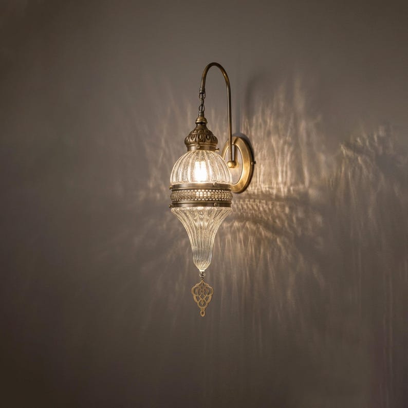 May include: A wall-mounted lamp featuring a ribbed, clear glass shade with a teardrop shape. The fixture has an antique brass-toned metal arm and a decorative metal band around the center. A small, ornate metal pendant hangs from the bottom tip.