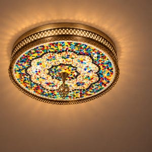 May include: A round flush mount ceiling light fixture featuring a multi-colored mosaic glass shade with a geometric pattern. The fixture has a metallic bronze-toned frame with a decorative filigree edge and a central metal finial.