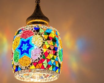 May include: A hanging pendant lamp featuring a cylindrical glass shade covered in a colorful mosaic of small, multi-shaped tiles. The design includes blue star patterns and circular motifs in red, yellow, and green, suspended by a bronze-toned metal cap.