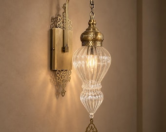 May include: A wall-mounted lamp featuring an antique brass finish backplate with intricate cut-out metalwork. A curved arm supports a clear, ribbed glass shade with a teardrop shape, topped by a decorative brass cap and a dangling metal finial.