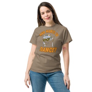 Op de afbeelding: Een taupekleurig T-shirt met korte mouwen en een afbeelding van een kat met een zonnebril die een dab-pose aanneemt. De tekst "WHO WANTS TO DANCE?" staat in een oranje, vetgedrukt en verweerd lettertype boven en onder de illustratie.