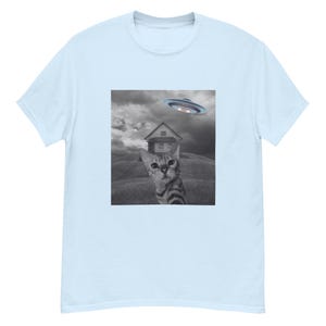 Op de afbeelding: Een zwart-witfoto van een kat die omhoog kijkt naar een vliegende schotel. De kat zit voor een klein huis. De foto is gedrukt op een lichtblauw T-shirt.