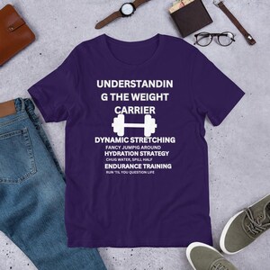 Op de afbeelding: Paars T-shirt met witte tekst die "Understanding the Weight Carrier" luidt en een halter afbeelding bevat. De tekst onder de afbeelding luidt "Dynamic Stretching, Fancy Jumping Around, Hydration Strategy, Chug Water, Spill Half, Endurance Training, Run 'Til You Question Life."