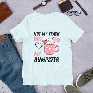 Op de afbeelding: Lichtblauw T-shirt met een afbeelding van een kat in een mok met harten en de tekst "NOT MY TRASH NOT MY DUMPSTER".