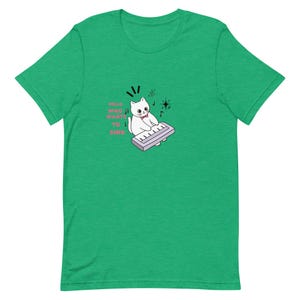 Op de afbeelding: Een felgroen T-shirt met korte mouwen en een afbeelding van een witte kat met een roze strikje die op een lichtpaars keyboard speelt. De tekst HELLO WHO WANTS TO SING staat in het roze naast de kat, vergezeld door muzikale noot-pictogrammen.