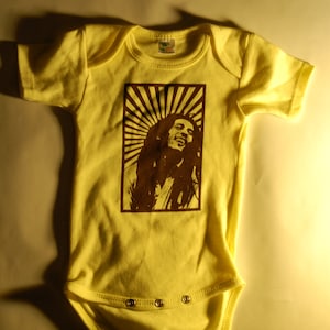 Puede incluir: Un body amarillo para bebé con una silueta marrón de Bob Marley en la parte delantera. La silueta está rodeada por un diseño de sol marrón.